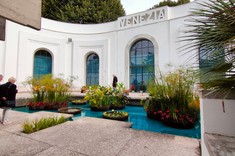 Wnętrze ogrodu otoczonego łukiem białych ścian budynku. W centralnej części ogrodu oczko wodne z kępami zieleni. W górnej części ściany widoczny napis VENEZIA.