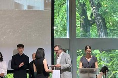 Kolorowe zdjęcie przedstawia wręczenie nagrody. Na pierwszym planie pani Regina Otto ubrana na czarno odbiera nagrodę od członka jury konkursu, mężczyzny ubranego w jasny garnitur. W tle duże przeszklenie i za nim ściana zieleni. 