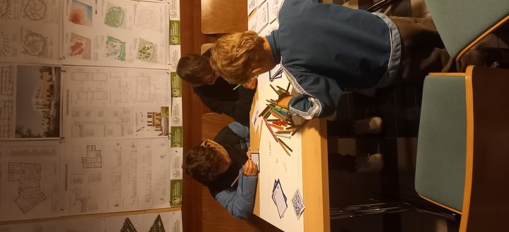 Zdjęcie wewnątrz budynku, na ścianach widoczne plansze z pracami studentów, przy stole siedzi trójka dzieci rysujących. 