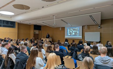 Wewnątrz sali z jasnym drewnopodobnym wykończeniem ścian i białym sufitem zgromadzonych jest kilkadziesiąt osób podczas wykładu. Na zdjęciu w oddaleniu widoczny jest ekran z prezentacją i prelegent, wszyscy słuchacze siedzą.