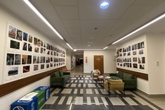 W pomieszczeniu z jasnymi ścianami i sufitem na ścianach po prawej i lewej stronie zawieszone duże plansze z wyeksponowanymi fotografiami w stonowanych barwach. Pod ścianami stoją fotele i kanapy.