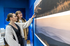 Przed planszami z fotografiami pejzaży górskich stoją dwie kobiety. Uśmiechają się. Jedna z nich wskazuje drugiej jakiś fragment zdjęcia.