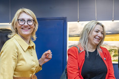 Dwie kobiety stoją obok siebie i uśmiechają się do fotografii. Obie z jasnym włosami, jedna w żółtej sukience, druga w czerwonej marynarce.