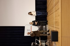 Zdjęcie przedstawia trzy osoby stojące na scenie podczas wydarzenia. Po lewej stronie, znajduje się mężczyzna w brązowej marynarce, który mówi do mikrofonu. Obok niego stoją dwie kobiety – jedna w czarnej bluzce i ciemnych spodniach, druga w jasnej koszuli i jeansach.