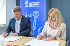 Od lewej rektor, prof. P. Koszelnik, dr. B. Szafrańska. Dwie osoby, kobieta i mężczyzna siedzą przy stole i podpisują umowę. Oboje elegancko ubrani, mężczyzna w ciemną marynarkę, kobieta na jasno. W tle niebieski baner z białym napisem Politechnika Rzeszowska.