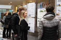 Na zdjęciu grupa rozmawiających ze sobą studentów. W tle stoi wystawa prac konkursowych: prostokątne plansze zawieszone na metalowych siatkach,