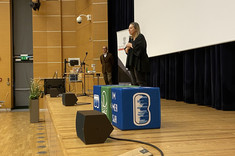 Na zdjęciu widzimy scenę w dużej sali audytoryjnej. Na środku sceny stoi kobieta ubrana na czarno, przemawiająca do publiczności. W tle widoczny jest mężczyzna w brązowej marynarce. Na scenie znajdują się także sprzęty techniczne – mikrofony, komputer oraz ekran projekcyjny, który zajmuje dużą część prawej strony kadru. Na podeście znajdują się niebieskie i zielone kostki z logotypami.