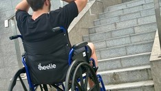 Młody mężczyzna, ubrany na czarno, siedzi na wózku inwalidzkim przed schodami, trzyma ręce wzniesione ku górze.