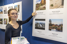 Zbliżenie na młoda kobietę z jasnymi włosami i w okularach. Kobieta wskazuje ręką na zdjęcie, w drugiej ręce trzyma nagrodę.