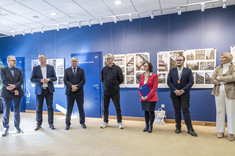 W jasno oświetlonym wnętrzu, z ciemnoniebieskimi ścianami, na tle plansz wywieszonych w rzędzie na ścianie, stoi siedem elegancko ubranych osób.