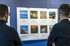 Na białej planszy umieszone osiem kolorowych zdjęć. Plansza zawieszona na ciemno niebieskiej ścianie. Dwóch mężczyzn ubranych na czarno przygląda się planszy ze zdjęciami.  