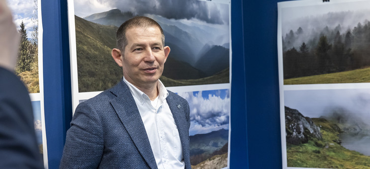 Na pierwszym planie stoi autor wystawy, mężczyzna w ciemnej marynarce.  W tle na granatowej ścianie zawieszone plansze z dużymi kolorowymi fotografiami pejzaży górskich.