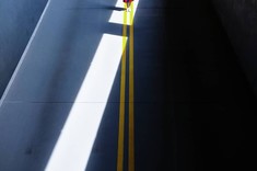 Betonowy tunel, po przekątnej wyraźna smuga jasnego światła. Smugę przecina żółta podwójna linia drogowa. Na linii stoi kobieta w kolorowej spódnicy.