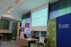 W lekko przyciemnionym pomieszczeniu po środku wysoko zawieszony jasny ekran, przed nim przy stole siedzi prelegentka. W tle kilka wysokich na około 2 metry plansz w zielonym lub niebieskim kolorze z białymi napisami.