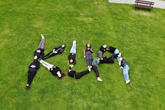 Zdjęcie wykonane z wysokości kilku metrów. Na zielonej trawie leży kilka osób , których sylwetki tworzą napis KUA.