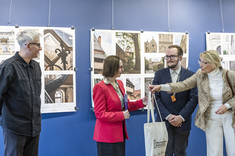 Kobieta ubrana na jasno wręcza kobiecie w czerwonej marynarce prezent. Obok nich stoją dwaj mężczyźni ubrani na ciemno.