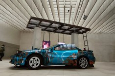 W jasnym, dobrze doświetlonym wnętrzu w jednolitej jasnoszarej kolorystce ścian, podłogi i sufitu stoi sportowy samochód w jaskrawo niebieskim kolorze.