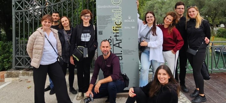 Grupa dziecięciu osób, swobodnie rozmieszczonych wokół pionowego banneru reklamowego pozuje do wspólnego zdjęcia. Wszyscy się uśmiechają. Na banerze, na szarym tle umieszczony napis w pionie Artificial Collective i czerwony znak biennale.  W tle wysokie drzewo.