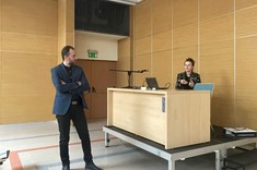 W jasnym wnętrzu sali wykładowej ze ścianami z drewnopodobną wykładziną na podwyższeniu za stołem siedzi prelegentka prowadząca wykład, przed stołem stoi mężczyzna słuchający wykładu. 