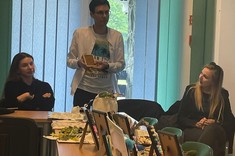 W pomieszczeniu na długim stole wystawione różne prace plastyczne. Za stołem stoi i tłumaczy coś mężczyzna, słuchają go dwie kobiety siedzące za stołem.