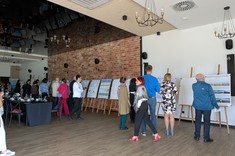 Pomieszczeniu z białymi ścianami i fragmentami z ceglanym wykończeniem. Ujęcie wykonane z oddalenia na grupę osób podczas przerwy w obradach. Liczne osoby analizują plakaty naukowe ustawione w linii pod ścianą. 