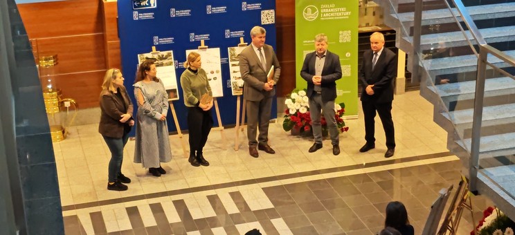 Zdjęcie wykonane z góry. Na tle ściany ustawione dwie duże płaszczyzny: niebieska i zielona, obie z białymi napisami. Przed płaszczyznami stoi w jednej linii sześć osób. Trzy kobiety po lewej stronie i trzech mężczyzn po prawej.