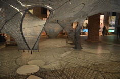 Nastrojowo oświetlone wnętrze. Po środku zawieszona duża przestrzenna rzeźba z szarej siatki, przez którą przebija delikatnie światło i rzuca na podłogę delikatny wzorzysty cień.