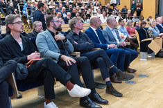 Duża grupa osób siedzących w rzędach podczas wykładu. Na pierwszym planie grupa pięciu mężczyzn.