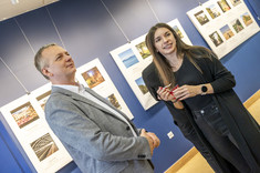 Na pierwszym palnie mężczyzna w jasnej marynarce i kobieta ubrana na czarno uśmiechają się do zdjęcia.  W tle plansze ze zdjęciami.