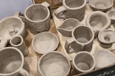 Zbliżenie na kubki i miski ulepione z gliny. Wszystkie przedmioty w stanie surowym w szarym kolorze. 