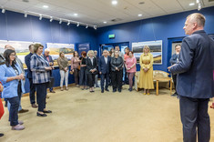 Ujęcie pomieszczenia, w którym w kilku grupach stoją uczestnicy wystawy. Wszyscy zwróceni w jednym kierunku, słuchają przemawiającego mężczyzny, widocznego po prawej stronie zdjęcia. W tle na granatowej ścianie zawieszone plansze z dużymi kolorowymi 