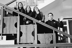 Zdjęcie czarno białe. Na schodach za drewnianą balustradą stoi na różnych wysokościach czwórka młodych osób, trzy kobiety i jeden mężczyzna. Wszyscy spoglądają prosto w obiektyw i uśmiechają się.