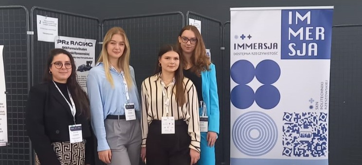 We wnętrzu, na tle ciemnoszarej ściany cztery młode kobiety. Wszystkie są ubrane elegancko w kolorach czarnym, beżowym i niebieskim. Wszystkie mają długie włosy. Po prawej stronie poster – biała płaszczyzna z napisem Immersja i wyraźnymi grafikami w kolorze ciemnoniebieskim.  