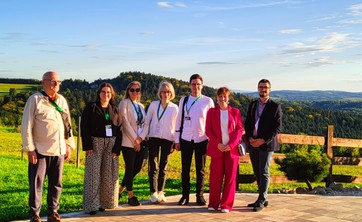 Grupa siedmiu osób pozuje do zdjęcia na tle naturalnego krajobrazu. Wszyscy są uśmiechnięci. W tle zielone wzgórza pokryte lasami i niebieskie niebo.