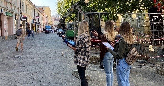 Widok na ulicę- deptak miejski w porze letniej. Środek dnia, kilkoro przechodniów. Po prawej grupa drzew, po lewej fasady historycznych niewysokich kamienic.  Na środku ulicy trzy studentki prowadzą pomiary akustyczne.