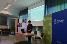 W lekko przyciemnionym pomieszczeniu po środku wysoko zawieszony jasny ekran, przed nim, przy stole stoi prelegentka. W tle kilka wysokich na około 2 metry plansz w zielonym lub niebieskim kolorze z białymi napisami.