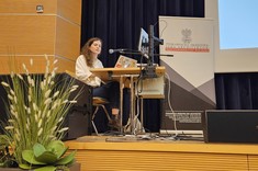 Na zdjęciu widzimy kobietę z długimi, falowanymi włosami, ubraną w jasną koszulę i jeansy, która siedzi przy biurku na scenie i prowadzi prezentację. Przed nią znajduje się laptop oraz sprzęt multimedialny, w tym mikrofon i monitor. Obok niej stoi baner. Na pierwszym planie widoczne są również ozdobne rośliny doniczkowe. Tło stanowi ciemna zasłona sceniczna i fragment dużego ekranu projekcyjnego.