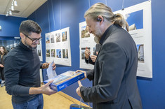 Mężczyzna w ciemnej marynarce wręcza nagrodę mężczyźnie w ciemnym golfie.  W tle plansze ze zdjęciami.