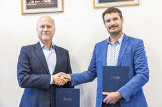 Dwóch mężczyzn elegancko ubranych stoi na tle białej ściany.  W rękach trzymają okładki z dokumentami. Podają sobie ręce i uśmiechają się.