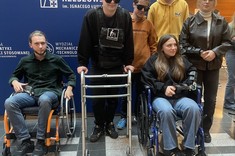 Zdjęcie wewnątrz budynku przedstawiające grupę studentów podczas zajęć. Studenci na wózkach  inwalidzkich, wyposażeni w symulator starości, okulary symulujące wady wzroku. 