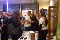 Grupa osób stoi przy stole z ulotkami i okularami. Studentka w białej koszulce gestykuluje, tłumacząc coś zainteresowanym. Obok stoi wózek inwalidzki. W tle widoczne są stoiska innych wydziałów oraz świetlówka w kształcie miecza świetlnego.