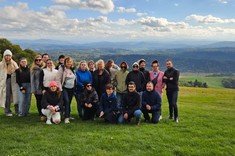 Zdjęcie grupowe na tle panoramy gór. Bardzo dobrze widoczna panorama gór – łagodne pofałdowania, kolorystyka wczesnojesienna. Niebo jest lekko zachmurzone. Na pierwszym planie stoi grupa około 20 uczestników warsztatów. Wszyscy ubrani na sportowo w kolorach niebieskim, czarnym i beżowym.