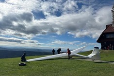 Zdjęcie pięknej panoramy gór. Niebo niebieskie z niewielkimi grupami białych chmur. Na pierwszym planie, na zielonym trawniku biały szybowiec przygotowywany przez kilka osób do lotu. 