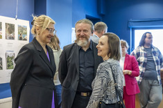 Zdjęcie grupy osób na tle wystawy. Na pierwszym planie dwie kobiety i mężczyzna podczas rozmowy, uśmiechają się.