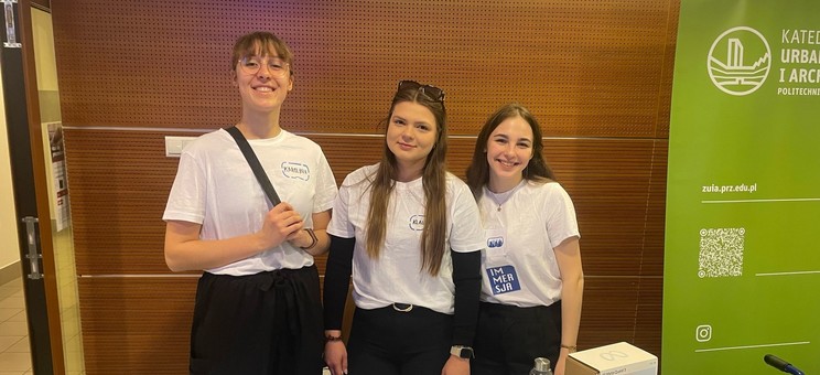 Na zdjęciu trzy uśmiechnięte studentki stoją za stołem przykrytym białym obrusem, na którym znajdują się ulotki, okulary i gadżety. Z prawej strony stoi zielony baner Katedry Urbanistyki i Architektury.