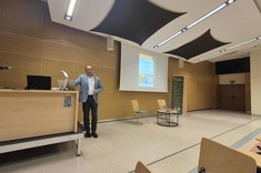Pomieszczenie z białym sufitem i drewnopodobnym wykończeniem ścian. Po środku przy podwyższeniu stoi mężczyzna podczas prowadzenia wykładu. W tle ekran z wyświetlonym slajdem.  