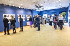 Wnętrze z ciemno niebieskimi ścianami. Na ścianach zawieszone białe plansze z kolorowymi zdjęciami. We wnętrzu, w kilku luźnych grupach, zgromadzone osoby odwiedzjące wystawę.