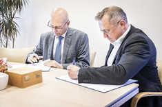 Na zdjęciu w jasnym pomieszczeniu dwóch mężczyzn w garniturach podpisuje dokumenty.