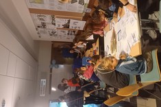 Zdjęcie przedstawia grupę ludzi znajdujących się w korytarzu w budynku. Na ścianach znajduje się wystawa prac studentów. Dzieci siedzą przy stołach, rysują, naklejają, biorą udział w zabawach sensorycznych – dotykowych. 