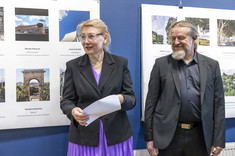 Kobieta i mężczyzna w ciemnych marynarkach stoją na tle plansz ze zdjęciami. Kobieta trzyma w ręku białą kopertę.